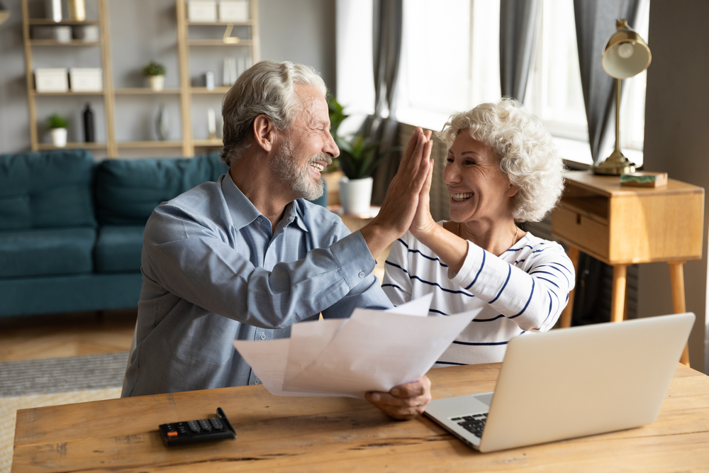 A senior couple give each other a high-five while looking at paperwork and a computer