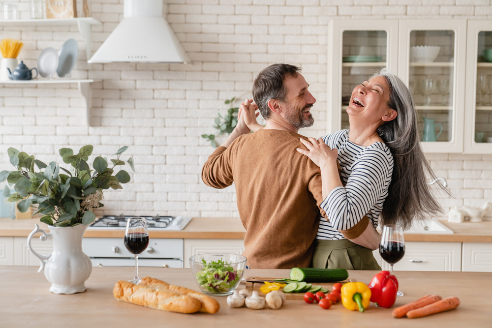 A senior man and a senior woman have a romantic spin in the kitchen