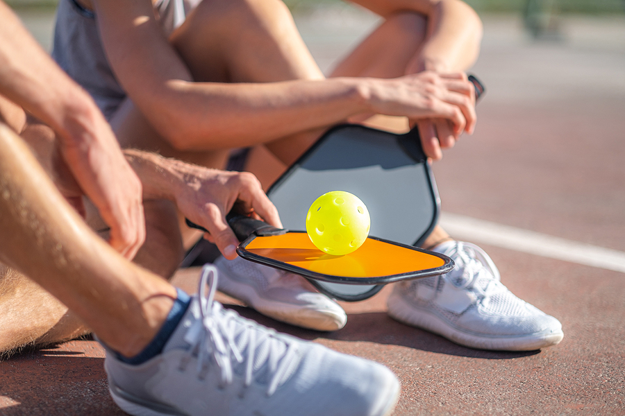 Two friends sit on the ground with pickleball paddles.