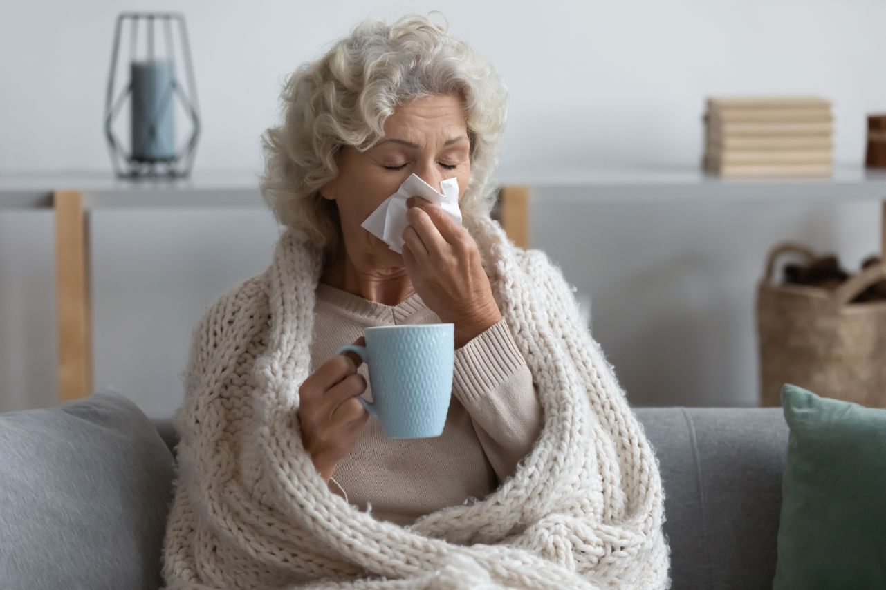 Senior sitting on the couch sick and blowing nose while holding a cup of tea.
