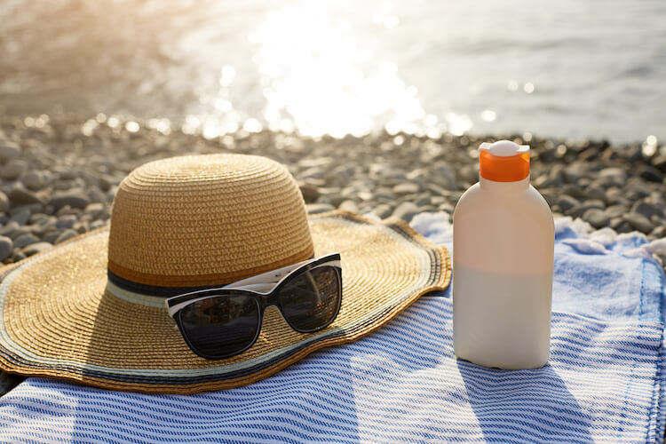Sunscreen bottle, a hat and sunglasses on a beach towel with sea shore on background.