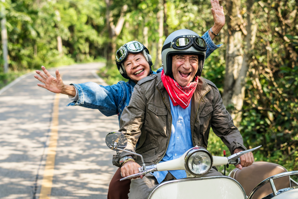 A senior woman and senior man ride a moped, laughing and wearing goggles and a helmet